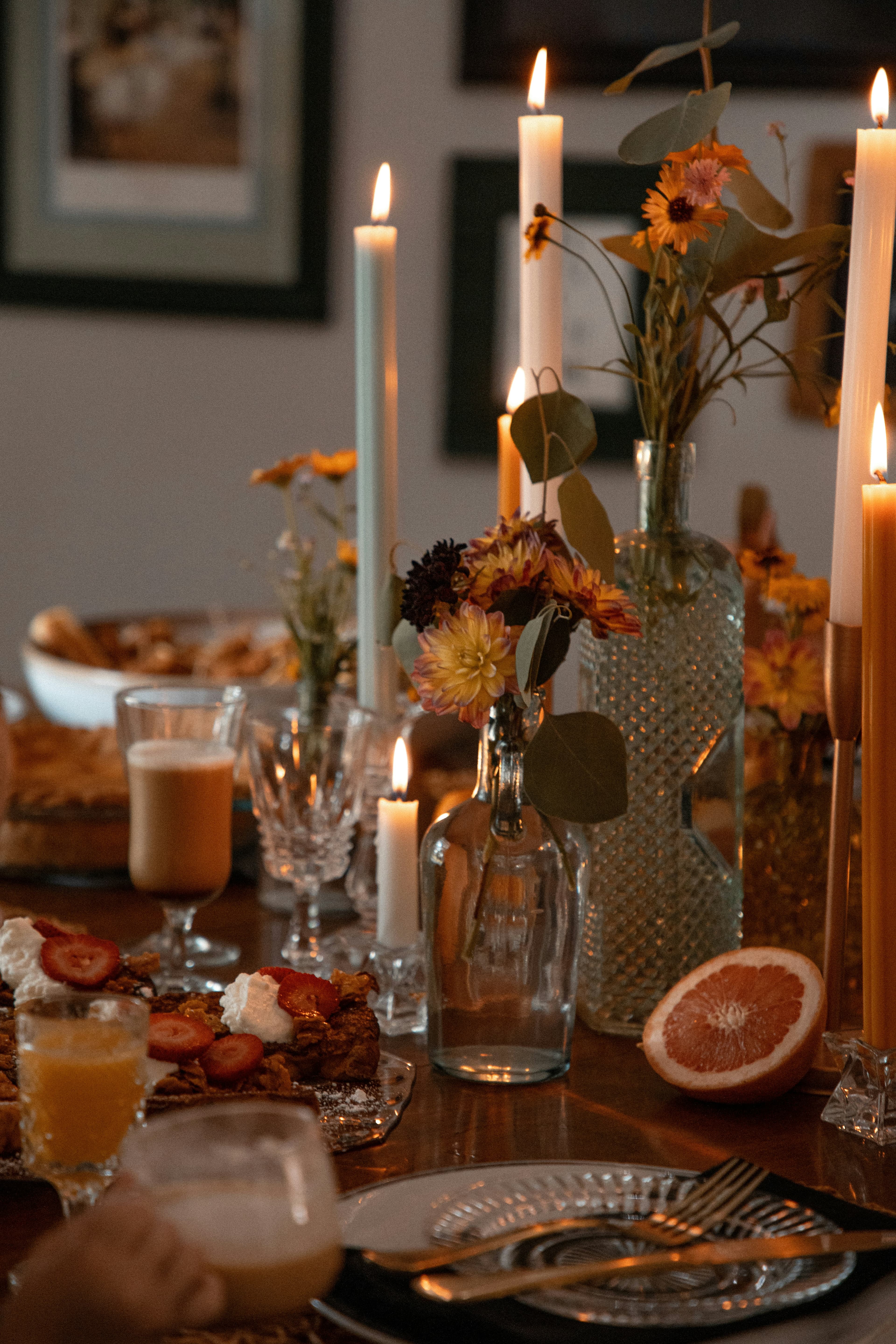 A warmly lit table with tall taper candles, flowers and pudding